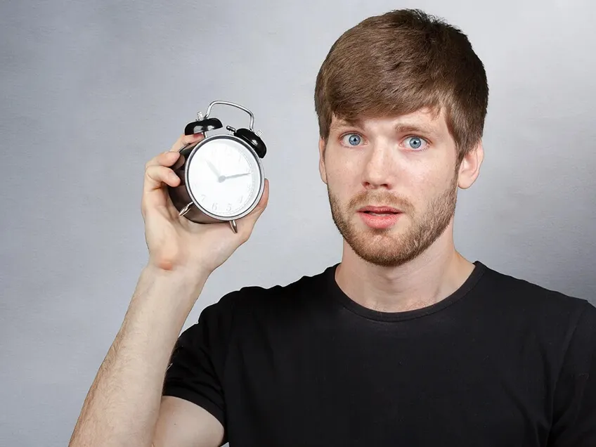 Man holding an alarm clock with a surprised expression, symbolizing time wasted in meetings.